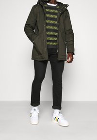 Olivengrøn polstret jakke med hætte, båret over en sort og neonmønstrede sweater, kombineret med sorte jeans og hvide sneakers.