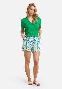 Grüne Kurzarmstrickjacke mit strukturiertem Muster und Knopfleiste; kombiniert mit weißen Shorts, die ein blaues und grünes Blumenmuster aufweisen.