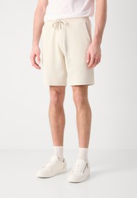 Pantaloni corto lavorati in maglia beige chiaro con vita elasticizzata e cordoncino, dotati di tasche laterali e finitura in tessuto testurizzato. Abbinati a scarpe bianche.