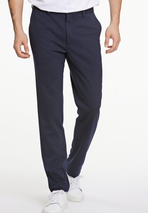 Marineblaue Slim-Fit-Hosen aus glattem Stoff, mit flacher Vorderseite, Seitentaschen und einem maßgeschneiderten Erscheinungsbild. Getragen mit weißen Sneakern.