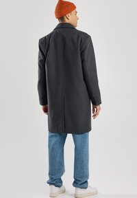 Cappotto di lana grigio scuro con colletto classico, spacco sul retro e polsini con bottoni, abbinato a jeans blu chiaro e un berretto lavorato a maglia arancione brillante.