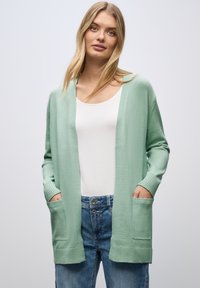 Světle zelený kardigan vyrobený z jemného pleteného materiálu, s uvolněným střihem, otevřeným předním dílem a dvěma předními kapsami. Spárován s bílým topem a džínami.