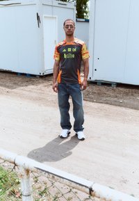 Grå jersey med gule og orange accenter, sorte jeans og hvide sneakers. Baggrunden har hvide shippingcontainere og en jordsti.