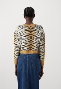 Pull en tricot avec un motif rayé tigre dans différentes nuances de crème, noir et or. Poignets et ourlet côtelés, porté sur une jupe en jean.