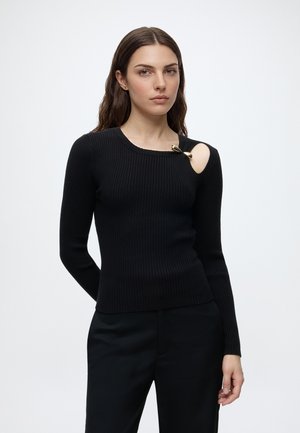 Jeune femme aux longs cheveux foncés porte un pull côtelé noir avec une épingle dorée sur la découpe à l'épaule et un pantalon noir sur un fond blanc uni.