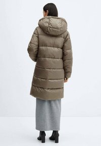 Manteau long matelassé vert olive avec des panneaux cousus horizontalement, une capuche et une coupe décontractée, associé à une longue jupe grise et des bottes noires.