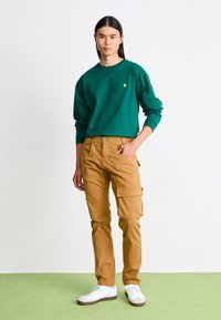 Grünes Langarmshirt mit kleinem Logo, kombiniert mit hellbraunen Cargo-Hosen mit Taschen. Weiße Sneaker runden das Outfit ab.