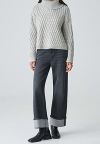 Femme portant un pull à col roulé gris clair en tricot torsadé, jean large sombre retroussé aux chevilles et bottines noires, debout sur un fond uni.