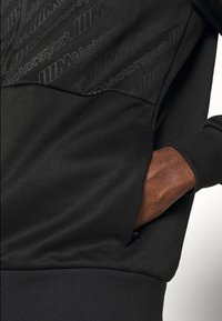 Svart sweatshirt med texturerad övre panel som har ett subtilt "M Motorsport" mönster, dragkedjeförsedd sidoficka och ribbade muddar.