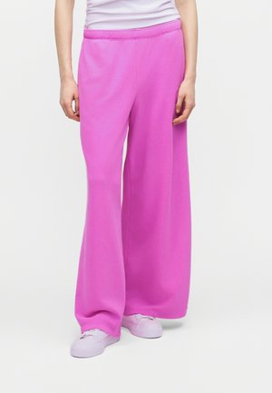 Person iført lyserøde vidde bukser, hvide sneakers og en hvid top stoppet ind i bukserne mod en ensartet lys baggrund.