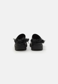 Svarta slip-on skor med fluffig trim, rund bakkant och platt gummisula, som visar upp en strukturerad yta och minimalistisk design.