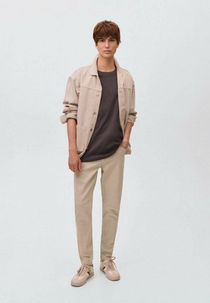 Veste en denim beige sur une chemise gris foncé, accompagnée de pantalons beige clair et de sneakers beige. Design simple avec des accents et des textures minimales.