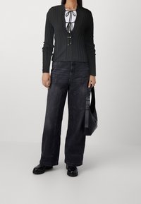 Schwarzer gerippter Cardigan mit tiefem V-Ausschnitt und Bindverschluss, kombiniert mit locker sitzenden dunkelblauen Jeans und einer schwarzen Umhängetasche.