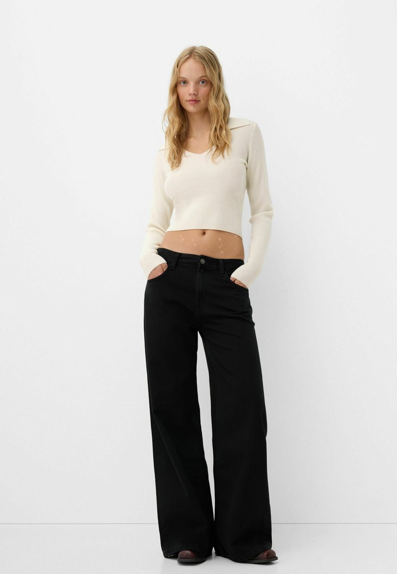 Top corto a costine crema con maniche lunghe e pantaloni neri a gamba larga. L'outfit presenta una chiusura con bottoni e tasche laterali.