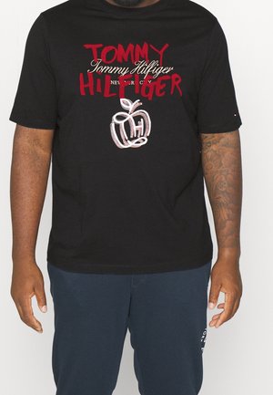 Schwarzes T-Shirt mit rotem und weißem "Tommy Hilfiger"-Schriftzug und stilisiertem Apfel-Logo, getragen von einer Person mit tätowiertem rechtem Arm und Marineblauer Hose.