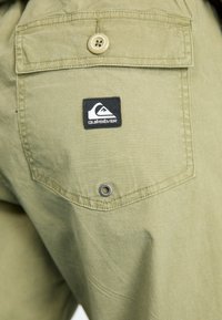Olive green shorts with a patched back pocket, featuring a button closure and a metal grommet detail. Includes a black Quiksilver logo patch.