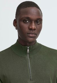 Grønn ribbestrikket genser med høy krage og halvglidelåsdetalj, tettsittende design og teksturert stoff.