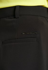 Musta räätälöity housut sileällä pinnalla, joissa on takatasku ja suorakulmainen metallikoriste, jossa on kaiverrettuna "Calvin Klein".