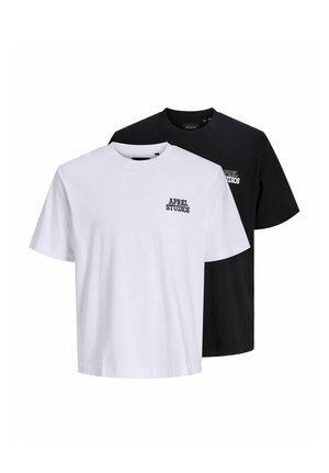 Uomo con berretto nero, T-shirt bianca a maniche corte con la scritta "APREL STUDIOS", pantaloni grigi larghi e scarpe beige, in piedi davanti a uno sfondo semplice.