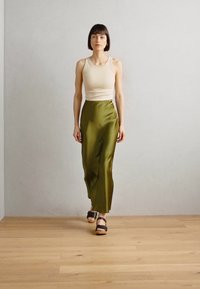 Beige tank top yhdistetty pitkään, valuvaan oliivinvihreään satiinishortsiin. Pukuun kuuluu litteät mustat sandaalit ja vaalean puun sävyinen lattia taustalla.