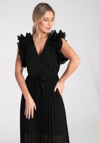 Schwarzes Kleid mit Rüschensleeves in Schichten, Wickeldesign und plissiertem Rock. Mit einem Bindegürtel in der Taille ausgestattet. Glatte Textur.