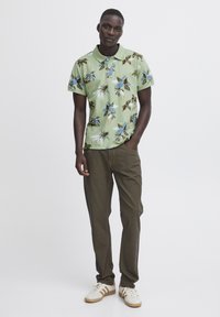 Grønn kortermet polo skjorte med blomsterprint i blått og brunt, kombinert med olivengrønne bukser og hvite sneakers med beige detaljer.
