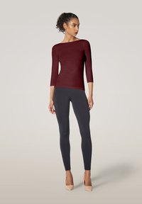 Bordeauxfarbener eng anliegender Oberteil mit drei Viertelärmeln, kombiniert mit schwarzen Leggings und nude-farbenen High Heels, vor einem neutralen Hintergrund.
