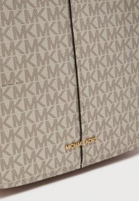 Μπεζ τσάντα καμβά με επαναλαμβανόμενο μοτίβο λογότυπου "MK", ολοκληρωμένη με επιχρυσωμένη πλακέτα "MICHAEL KORS" στο κάτω μέρος.