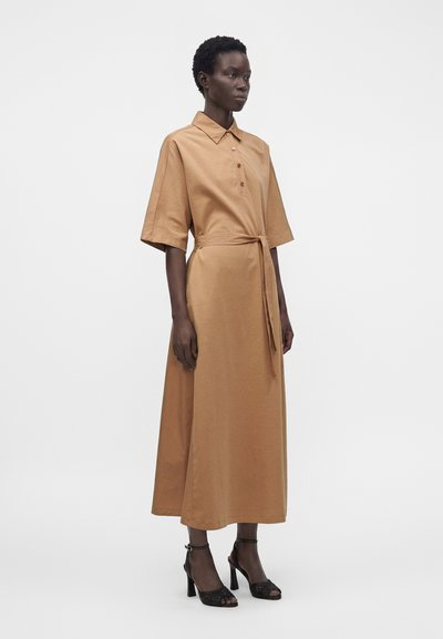 Femme portant une robe midi beige avec col, patte de boutonnage, manches mi-longues, ceinture à la taille et sandales à talons noires à bouts ouverts.