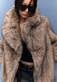 Cappotto in pelliccia sintetica in tonalità di marrone con un colletto voluttuoso e finitura testurizzata. Indossato sopra un top nero, abbinato a jeans denim scuri.