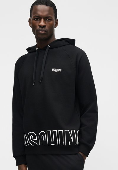 Mann iført svart Moschino Active-hettegenser med store hvite bokstaver nær kanten og liten logo på brystet, stående mot en ensfarget bakgrunn.
