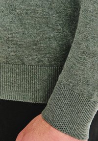 Zielony sweter z dzianiny, który charakteryzuje się teksturowanym wykończeniem, ściągaczami przy mankietach i u dołu. Materiał wydaje się miękki i lekki, z gładką, jednolitą powierzchnią.