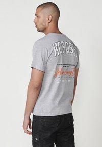 Light gray cotton T-shirt featuring bold black "KOROSHOP" text on the back, with additional branding and text in orange and black.