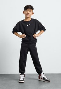 Črn Nike dukserica z belim logotipom, v kombinaciji s črnimi trenirkami in črnimi/rdečimi visoko-top majicami. Gladek material, sproščen kroj in rebrasti manšeti.