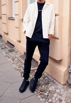 Personne appuyée contre un mur beige portant une veste blanche, un t-shirt noir avec un motif graphique, un pantalon noir et des baskets noires sur un trottoir en pavés.