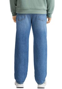 Persona che indossa jeans dritti blu, sneaker bianche e una felpa verde, vista di spalle su uno sfondo bianco.