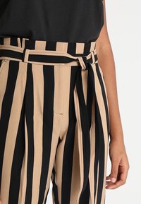 Beige en zwarte gestreepte high-waisted broek met een strikriem, voorzien van een gladde textuur en zijzakken, met verticale strepen.