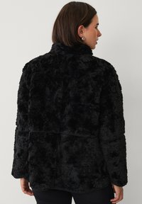 Sort faux fur jakke med høj krave, lange ærmer og en struktureret overflade. Ingen synlige lukninger eller mønstre.
