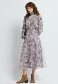 Mujer con cabello rizado vistiendo un vestido floral de cuello alto con mangas largas y botas marrones hasta el tobillo, de pie contra un fondo blanco liso.