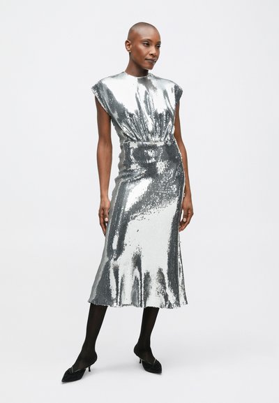 Sieviete, tērpusies bezpiekrītē, sudraba mirdzošā midi kleitā ar melnajām zeķubiksēm un melnajām norādītajām papēžiem, stāv uz balta fona.