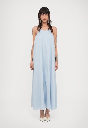 Kvinne med langt mørkt hår og tatoveringer, iført en lys blå ermeløs maxikjole og hvite spisse hæler, stående mot en ensfarget hvit bakgrunn.