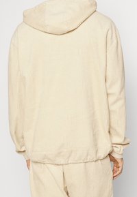 Beige kordfloddrad hoodie med ribbat mönster. Har en justerbar huva och elastiska ärmmuddar. Visad bakifrån, med en avslappnad passform.