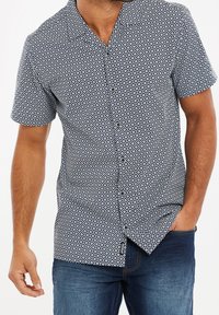 Chemise à manches courtes avec boutons, en tissu noir et blanc à motifs, présentant un design en losanges, un col et une coupe décontractée. Jean bleu visible.