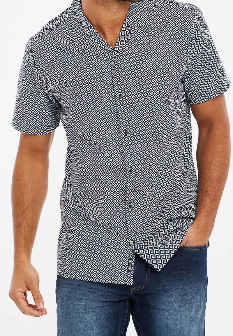 Chemise à manches courtes avec boutons, en tissu noir et blanc à motifs, présentant un design en losanges, un col et une coupe décontractée. Jean bleu visible.