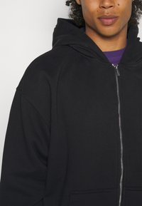 Czarna bluza z kapturem na zamek błyskawiczny, wykonana z mieszanki bawełny, z kapturem, ściągaczami przy mankietach i luźnym krojem. Zamek błyskawiczny biegnie pionowo wzdłuż przodu.