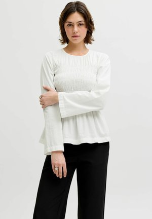 Lühikeste pruunide juustega naine, kes kannab valget pika varrukaga topi ja musti pükse, poseerib käed risti rinnal.