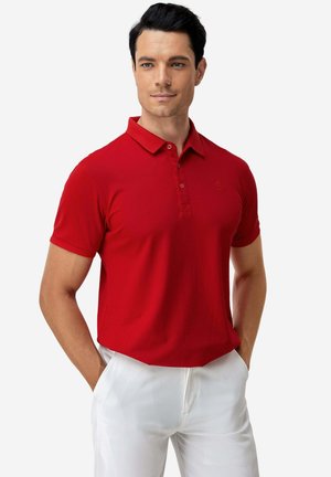 Mann mit dunklen Haaren, der ein rotes kurzärmeliges Poloshirt und weiße Hose trägt und mit den Händen in den Taschen vor einem einfarbigen Hintergrund steht.