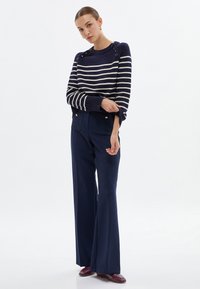 Marine gestreifter Pullover mit Knopfakzenten an den Schultern, kombiniert mit weiten Marine-Hosen, die goldfarbene Knöpfe und Fronttaschen haben.