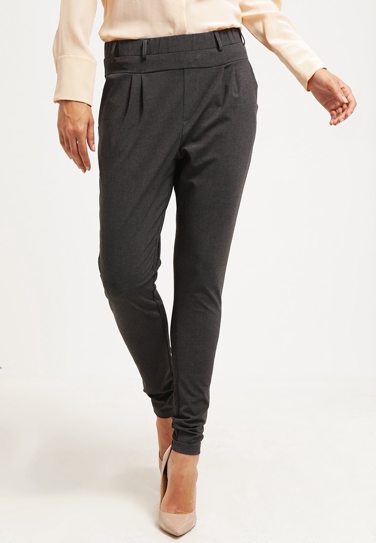 Femme portant un pantalon slim gris foncé et un chemisier beige, assortis à des chaussures à talons beiges, debout devant un fond uni.