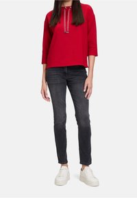 Červený svetr s třičtvrtinovými rukávy a stahovací šňůrkou u krku, v kombinaci s tmavými džíny a bílými teniskami. Pohodlný outfit na každodenní nošení.